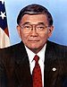 Older Japanese American man with glasses wearing a suit with a red tie with the US flag behind him