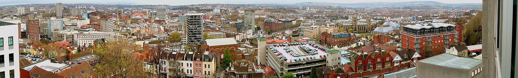 Een panoramisch uitzicht over een stadsbeeld van kantoorgebouwen, oude gebouwen, kerktorens en een parkeergarage.  In de verte zijn heuvels.
