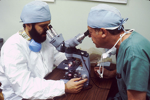 A pathologist examines a tissue section for evidence of cancerous cells while a surgeon observes.