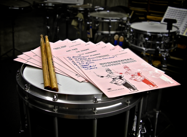 Rudimental Contest Series sheet music, perched atop a modern marching snare drum, with the cover featuring an illustration of Frank Arsenault Rudimental Contest Series portrait.png