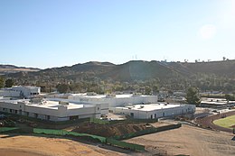 Elevated view of a cluster of low, white rectangular buildings with a line of hills in the near background.