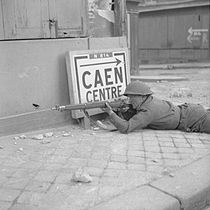 A soldier lies prone, rife at the ready, by a building in a city street. Beside him is a sign reading "Caen centre", pointing back the way he has come.