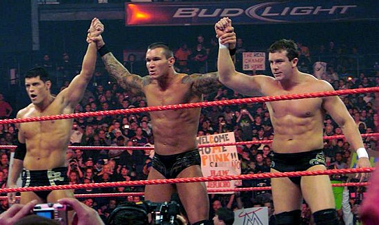 Three dark-haired men stand side-by-side in a wrestling ring with red ropes. The man in the center has tattoos on both his arms which are being raised in the air by the two men on either side of him. All three are wearing black wrestling tights.