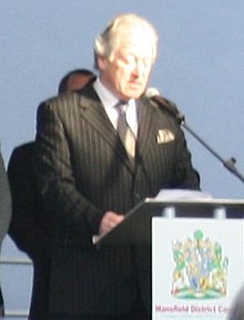 Silver haired man wearing a dark pinstripe suit on a raised platform giving a speech to spectators at the opening of a refurbished swimming pool