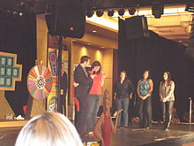 Marty Lublin interviewing a woman on a small stage. Three other women and a roulette-style wheel are also visible on the stage