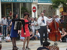 Photographie de 3 comédiens dans la rue : celle de gauche tient un accordéon, celui de droite une contrebasse (sur roulettes). Le public est assis ou debout derrière.