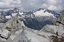 Roches gris clair angulaires au premier plan.  Derrière, une chaîne de montagnes grises plus foncées avec de la neige.  Celui de droite a un glacier important à son pied.