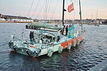 Un voilier dans le port, voiles baissées. La navigatrice lève les bras pour saluer.