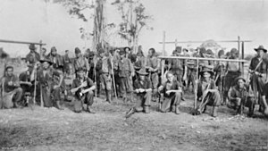 Foto hitam dan putih dari pasukan Australia, bersenjata dengan senapan, berpose dengan cara yang santai di depan sekelompok tahanan Jepang.