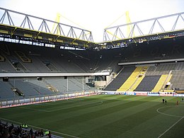 Two-tiered stands with grey seats, below the stands is a pitch with white markings