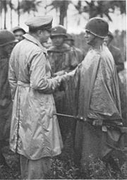 Officer in raincoat shakes hand of soldier wearing steel helmet and waterproof poncho while other similarly attired soldiers look on.