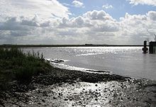 Muddy bank in the foreground before an expanse of water, with a concrete wall just visible on the right hand side of the water. In the distance is a line of low hills.