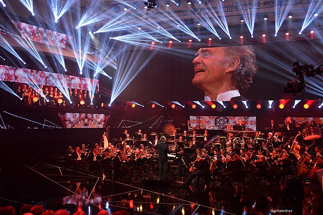 Large concert hall with an orchestra playing, and the conductor's face projected on a large screen over the stage