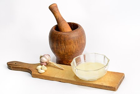 Mortar and pestle with cloves of garlic and a bowl of garlic sauce on a cutting board