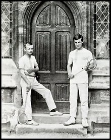 Two schoolboys holding racquets, standing on wooden steps either side of an arched wooden double door to a school building