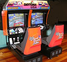 An arcade cabinet with two seats, two screens, and two steering wheels with pedals