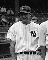 Shot vanuit een gemiddeld perspectief van honkbalspeler Lou Gehrig die lacht en een 'NY'-shirt en hoed draagt.