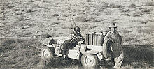 Man standing at the rear of a jeep which is heavily loaded with fuel cans and armed with twin guns at the front