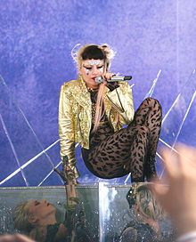 Gaga assise sur bocal de verre alors que deux femmes la regardent fixement tout en la tirant à l'intérieur du contenant.