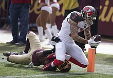 Een American football-speler vangt een bal terwijl een tegenstander op de grond deze probeert te stoppen.