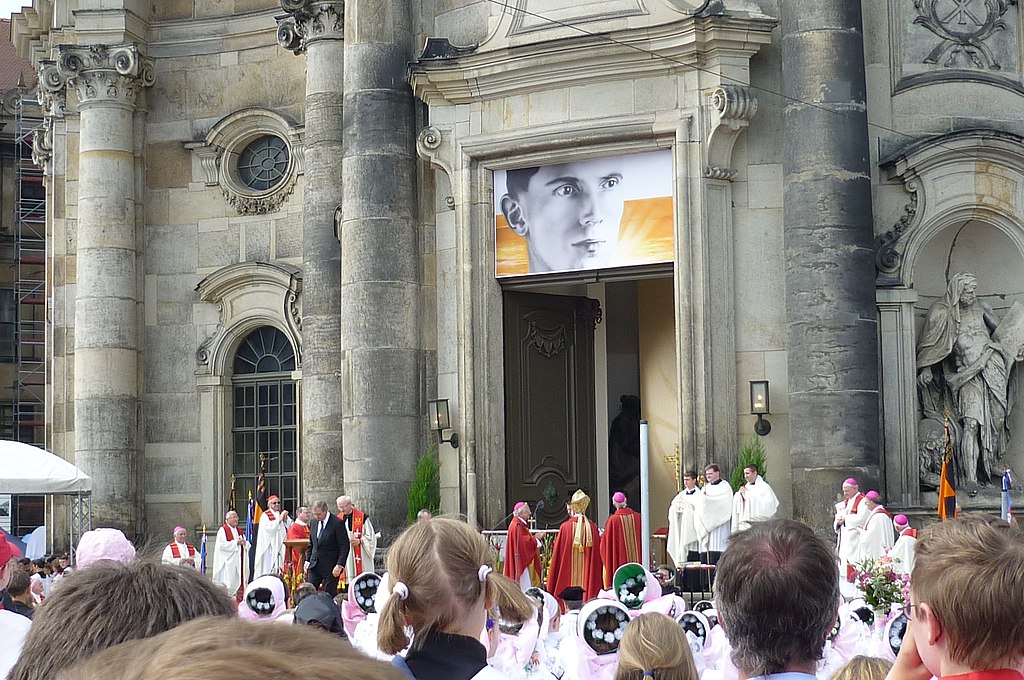 Photo de la façade d'un cathédrale avec plusieurs prélats, la foule et une grande photo d'un homme jeune