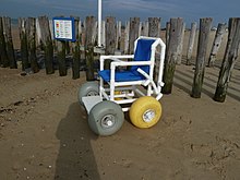 Ein Stuhl mit vier breiten, ballonartigen Rädern steht vor einem Zaun am Strand