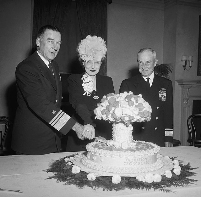 Admiral Blandy and his wife slice into a cake labeled Operation Crossroads, and shaped like a mushroom cloud, while Admiral Frank J. Lowry looks on, November 7, 1946.