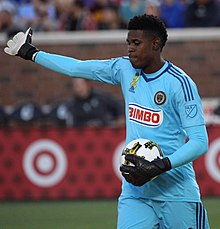 Andre Blake, wearing a blue goalkeeper jersey with the Philadelphia Union and Bimbo logos, holds a ball in his left glove with his right arm outstretched.