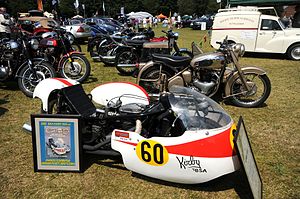 Low racing three wheeler motorcycle with sidecar combination wearing predominantly white with red streamlining on grass having a background of road going classic motorcycles and classic cars on a sunny day