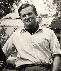 Viewed from the waist up: a stocky man wearing a pale-coloured button-up shirt with rolled-up sleeves, smoking a cigarette, looking directly at the camera, and leaning against the windshield of an amphibious vehicle.