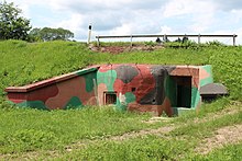 Pohled z východní strany na atypický bunkr vybudovaný pod silnicí. Pod komunikací je zatravněný svah silničního tělesa, z něhož vystupuje vstup do bunkru a střílna.