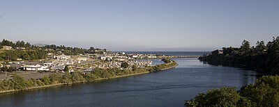 Um rio largo e suave flui pelo centro.  Sua margem direita é íngreme e arborizada, enquanto sua margem esquerda se desenvolve com muitos edifícios e estradas, e uma marina repleta de veleiros.  Os cais e o Oceano Pacífico são visíveis ao fundo, juntamente com um céu cinzento enevoado.