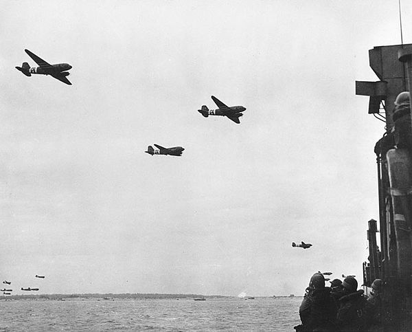 C-47 Skytrains with paratroops above a landing craft.