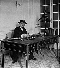 Weißer Mann mit ordentlichem Kaiserbart und Schnurrbart, sitzend an einem Schreibtisch, in einem Strohhut