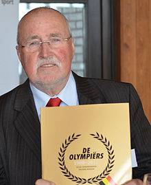 Farbfoto vom 75-jährigen Gaston Roelants im Anzug, in der Hand das Buch Olympiërs 1900-2012, das er als Vorsitzender des Vereins belgischer Olympiateilnehmer präsentiert.