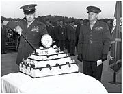 In front of a group of men, a man cuts a cake with a sword while a second man looks on