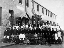 Posé, photo de groupe d'élèves et d'enseignants, vêtus de noir et blanc, à l'extérieur d'un immeuble en brique à Regina, en Saskatchewan