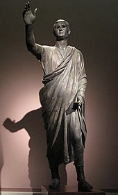 The Orator, c. 100 BC, an Etrusco-Roman statue of a Republican senator, wearing toga praetexta and senatorial shoes; compared to the voluminous, costly, impractical togas of the Imperial era, the Republican-era type is frugal and "skimpy" (exigua). L'Arringatore.jpg