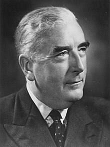 Head and shoulders portrait of 60-year-old man in 1940s business suit, wearing round horn-rimmed glasses and looking directly at camera.