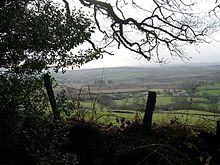 A view of hills and a patchwork of fields seen through a fence and tree.