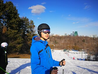 Felix, the Wikipedia user, is wearing ski goggles, a black ski helmet and his famous blue ski jacket in this photo. Behind him is a small ski hill.