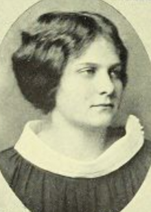 A young white woman with wavy hair parted center and dressed to nape; she is wearing a dark garment with a light-colored wide standing collar; in an oval frame