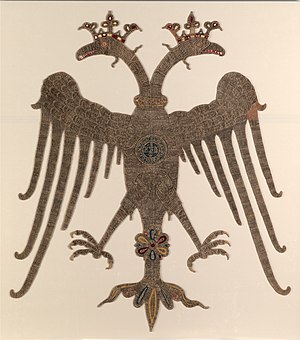 Photograph of a golden embroidered-double-headed eagle on an off-white background, with crowned heads and spread wings and legs, carrying a round medallion with Greek inscriptions on its breast.