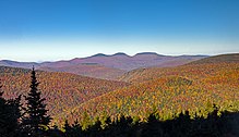 A landscape of forested mountains, in autumn color, underneath a blue sky, dominated by three rounded peaks in the distance with dark tops