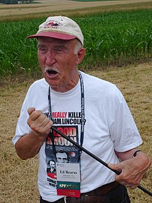 A white man wearing a t-shirt and ballcap is standing holding a staff and speaking.