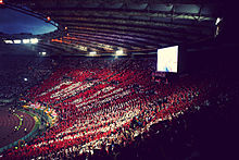 Stade rempli de spectateurs brandissant des cartons rouges et blancs formant un message.