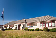 Gold Beach High School, Gold Beach. The schools is one-story, with a pitched metal roof. There is a lawn in front of the building featuring a flagpole and bench.