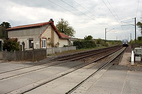 Pohled na malou budovu a nepoužívané nástupiště obsluhující dvě tratě s regionálním vlakem v pozadí, v zamračený den.