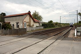 Vista de um pequeno prédio e plataformas desativadas servindo a dois trilhos, com um trem regional ao fundo, em um dia nublado.