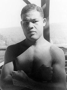 Photographie noir et blanc de Joe Louis qui pose torse nue, bras croisés et poings serrés.